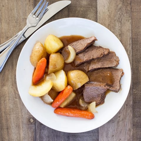 Sliced Beef Pot Roast served on a white plate with roasted root vegetables and a side of fresh herbs.