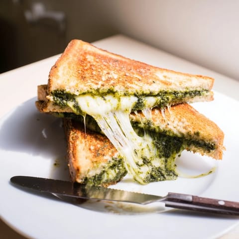 Golden, crispy slices of spinach pesto grilled cheese stacked on a plate, with a bowl of fresh spinach pesto beside it.